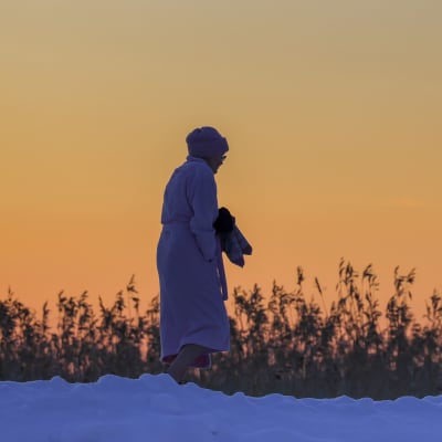 Henkilä aamutakki päällä kävelemässä avannosta pukukoppiin talvipakkasella auringonlaskun värjäten taivaan oranssiksi.