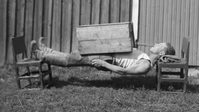 En man leker "ligga lik", där kroppen ska vara så styv att man ligger mellan två stolar med bara nacken och hälarna som stöd. Bilden är tagen 1932 i Esse.