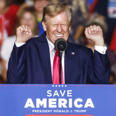 Donald Trump står vid ett talarpodium. Han håller upp händerna och grimaserar.