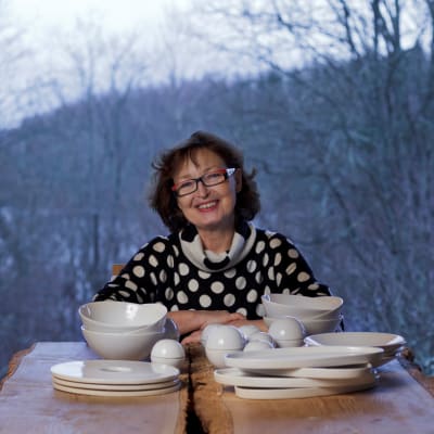 Kvinna med mörkt lockigt hår och glasögon sitter vid bord med keramik och ler.