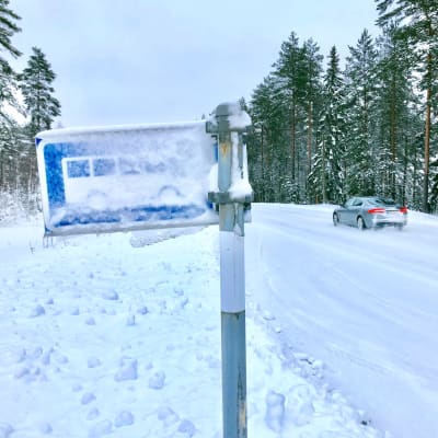 En nästan insnöad busskylt vid en busshållplats. Vinter och längs vägen kör en bil.