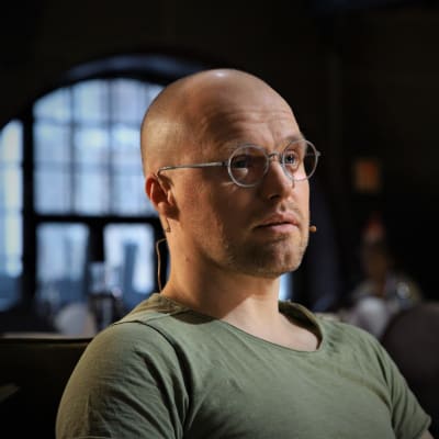 Kalju mies pyöreissä silmälaseissa ja vihreässä t-paidassa katsoo kameran ohi vakavana lähikuvassa. Tumma tausta, jossa näkyy ikkunasta häilyvää luonnonvaloa.