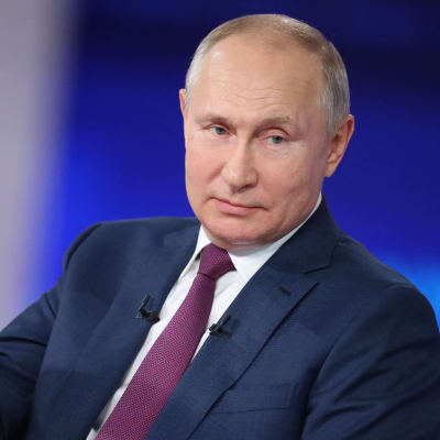 Vladimir Putin i närbild med huvudet på sned och höger handen utsträckt till sidan.