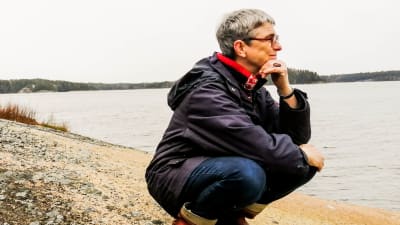 En medelålders kvinna på en strand tittar ut mot havet, ser glad och fundersam ut. 