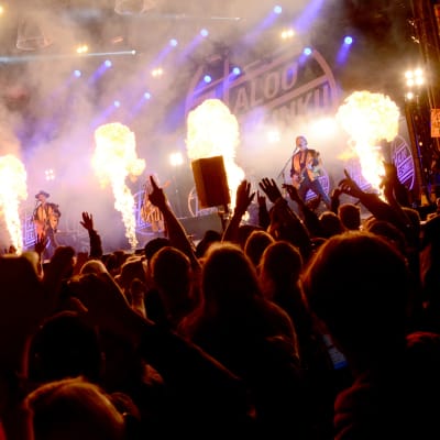 Rock-yhtye esiintyy lavalla, edessä yleisöä nauttii musiikista kädet ilmassa