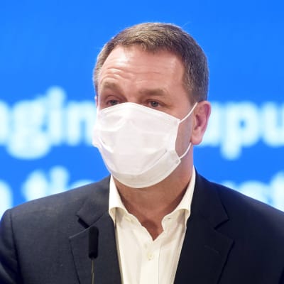 Jan Vapaavuori i munskydd på presskonferensen. Turkos bakgrund.