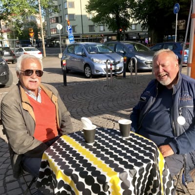 Två leende män sitter på ett torgcafé på Tölö torg.