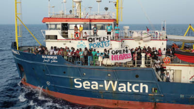En bild på båten Sea Watch med flyktingar ute på däcket med skyltar där det står "open ports" och "open hearts".