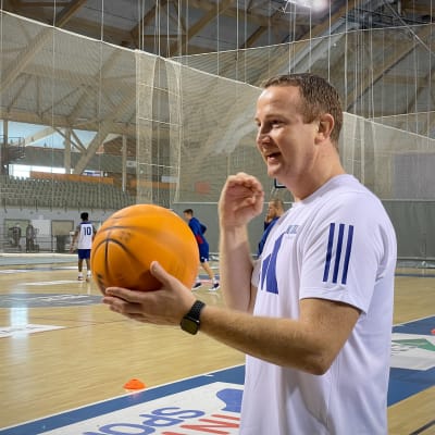 Kataja Basketin päävalmentaja Petri Virtanen pyörittää koripalloa käsissään, kun joukkue harjoittelee taustalla.