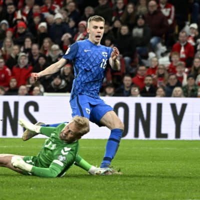 Suomen maajoukkueen peliasuun pukeutunut jalkapalloilija ohittaa pelikentällä maahan kaatunutta maalivahtia.