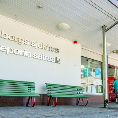 En äldre kvinna och en man påväg in i Raseborgs sjukhus. På byggnadens vägg står namnet på sjukhuset i silvriga bokstäver.