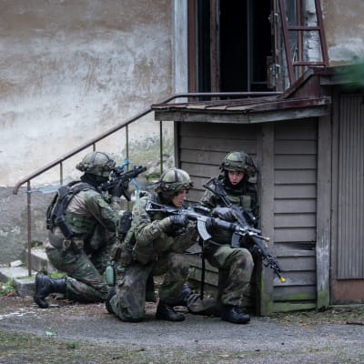 Fyra personer inom försvaret övar. De bär stridsutrustning med stormgevär och gömmer sig bakom en gårdsbyggnad.