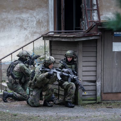 Fyra personer inom försvaret övar. De bär stridsutrustning med stormgevär och gömmer sig bakom en gårdsbyggnad.