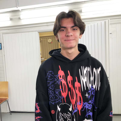 En ung man med vågigt mörkt hår och mörka ögonbryn tittar rakt in i kameran. Han har en svart munkjacka med färggranna symboler på. I bakgrunden finns en vit skolkorridor.