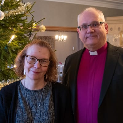 En kvinna och en man i anilinröd skjorta och prästkrage står leende framför en julgran.