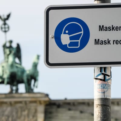 På en vit skylt står det att munskydd krävs - en bild av en person klädd i munskydd finns på skylten i blått. I bakgrunden syns ett monument där fyra hästar drar en vagn med en person i.