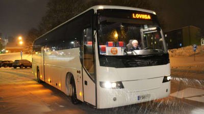 En vit buss står på en parkeringsplats. Inne i den annars tomma bussen sitter en äldre man som är chaufför. Uppe på bussen står Loviisa.