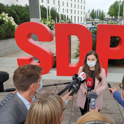 Sanna Marin intervjuas, flera personer står framför henne och räcker fram mikrofoner. I bakgrunden står det SDP med stora bokstäver.