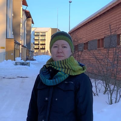 Kvinna i grön mössa och halsduk står med höghus i bakgrunden.