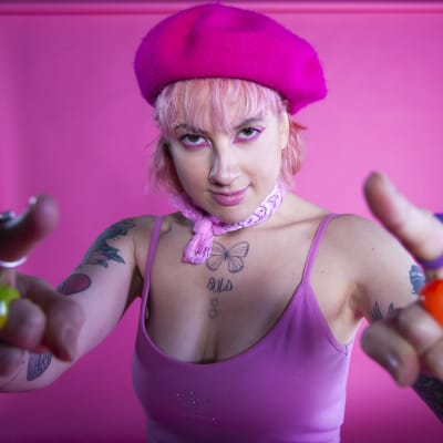 Närbild på Ronja Stanley som är klädd i rosa kläder och ser rakt in i kameran medan hon håller båda händerna uppe och pekar mot kameran.