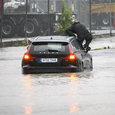 Någon klättrar ur bil på översvämmad väg. 