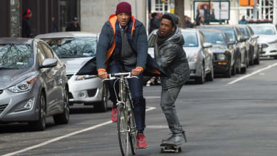 Howard cyklar medan Raffi/Tiden hänger fast vid hans pakethållade, balanserande på en skateboard.