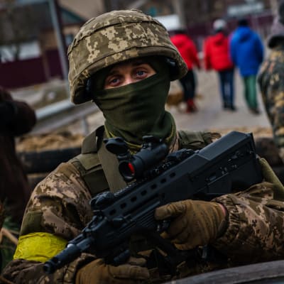 En soldat i full utrustning står och håller i ett vapen medan han tittar in i kameran. I bakgrunden fler soldater.