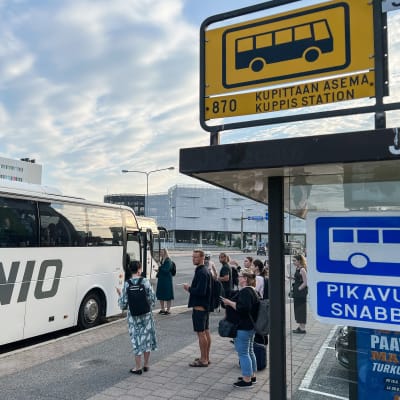 Ihmisiä odottaa bussipysäkillä nousua bussiin. Aamutaivaalla on pilviä. 