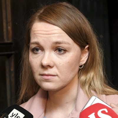 Katri Kulmuni, en ung kvinna med blont hår och en rosa jacka blir intervjuad framför en mörk vägg. I förgrunden syns två stycken stora mikrofoner och ett par ljudinspelare.