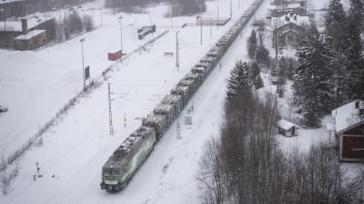 Ett tåg åker i snöväder, vid sidan om rälsen finns skog och byggnader och fält.