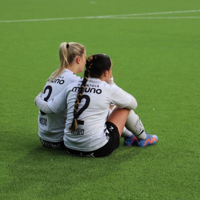 Två fotbollsspelare i vita skjortor och svarta byxor sitter på gräsmattan efter en match och håller armarna om varandra.