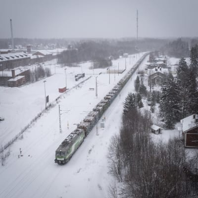 Ett tåg åker i snöväder, vid sidan om rälsen finns skog och byggnader och fält.