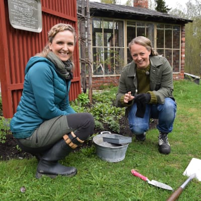Två leende kvinnor sitter på huk framför ett staket 