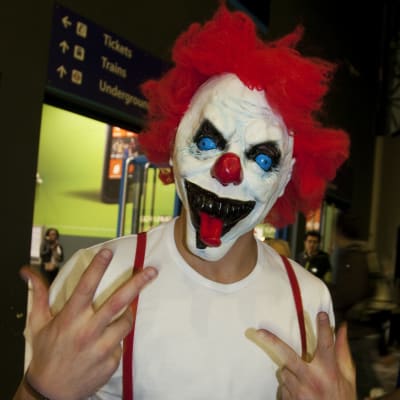 Person utklädd till clown, med en clownmask med rött hår och stora utstående blåa ögon.