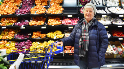 En äldre kvinna står med en kundvagn framför en hylla med lök i en matbutik.