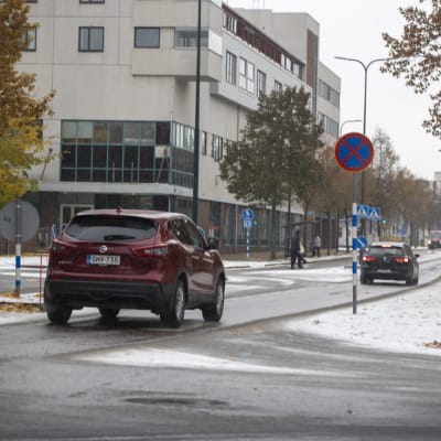 Kouvolan keskustassa autot ajavat Koulukatua, nainen on ylittämässä tietä jalan. Maassa on vähän lunta ja puissa on vielä keltaisia lehtiä