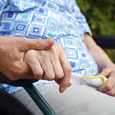 En äldre person i rullstol håller en vårdare i handen.