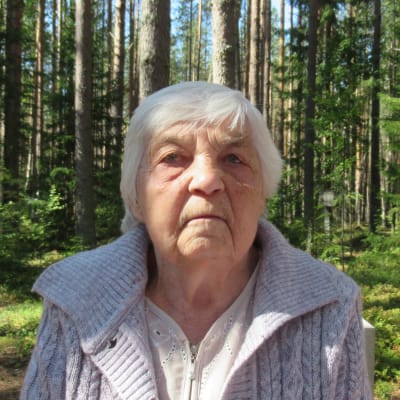 En äldre dam tittar rakt in i kameran. Bakom henne tät skog. 