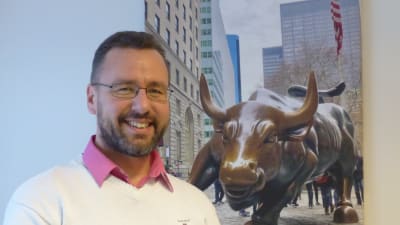 En man står framför en tavla som  är ett fotografi  av en staty i brons föreställande en tjur. Det är en bild tagen vid Wall Street i New York.