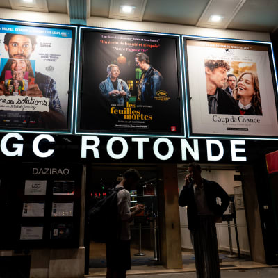 UGC Rotonde elokuvateatterin ovilla kaksi ihmistä puhelimillaan. Yläpuolellä kolmen elokuvan mainokset, joista keskimmäinen Aki Kaurismäen Kuolleet Lehdet ranskan kielisellä nimellä les feuilles mortes.