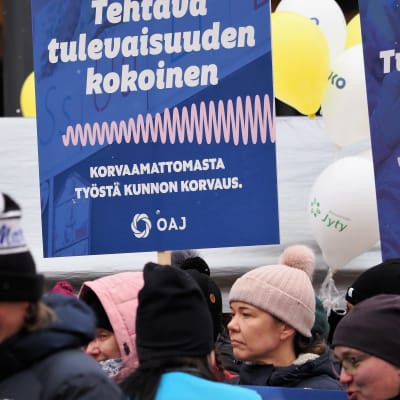 Mielenosoittajia ja taustalla sininen kyltti, jossa teksti Tehtäväkokonaisuuden kokoinen. Korvaamattomasta työstä kunnon korvaus OAJ.
