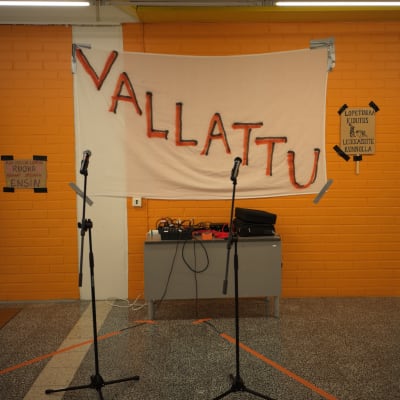 Kaksi mikrofonia, joiden takana on lakana, jossa lukee "Vallattu"