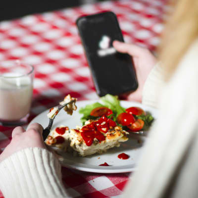 En person som sitter och äter makaronilåda. Ryggen på personen syns och i handen håller personen en telefon. Hen skrollar på sociala medier medan hen äter. 