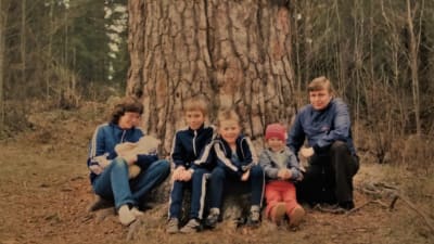 Perhe istuu ja poseeraa kameralle suuren männyn juurella: vasemmalla äiti sylissään vauva, keskellä kaksi kouluikäistä poikaa ja heitä nuorempi tyttö, oikealla isä.