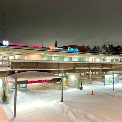 Sairaalan luminen etupiha ja pääsisäänkäynti. Sairaalan katolla värikkäitä kylttejä, jotka näkyvät yötaivasta vasten.