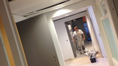 Målare i vit overall står i korridor i kontorslokal som renoveras, målarhink och -rulle på golvet.