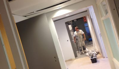 Målare i vit overall står i korridor i kontorslokal som renoveras, målarhink och -rulle på golvet.