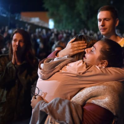 En kvinna omfamnar en person. I bakgrunden syns flera andra människor