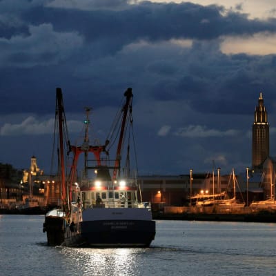 En fiskebåt utanför en hamn där andra båtar är förtöjda. Det är kväll och molnen i bakgrund är mörka och blåfärgade. Trålaren The Cornelis-Gert Jan har fått lämna hamnen i Le Havre efter att ha varit konfiskerad.