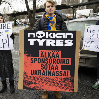 Mielenosoittajia kyltteineen Nokian Renkaiden tehtaan edustalla. Keskimmäisessä kyltissä lukee "Älkää sponsoroiko sotaa Ukrainassa". 